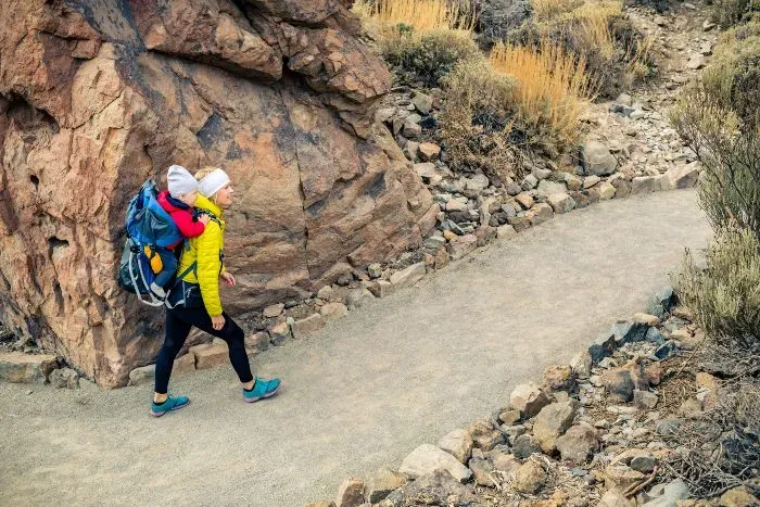 Madre cargando a su bebe, caminando sobre un sendero montañoso con el diseño de mochilas para transportar a pequeños.