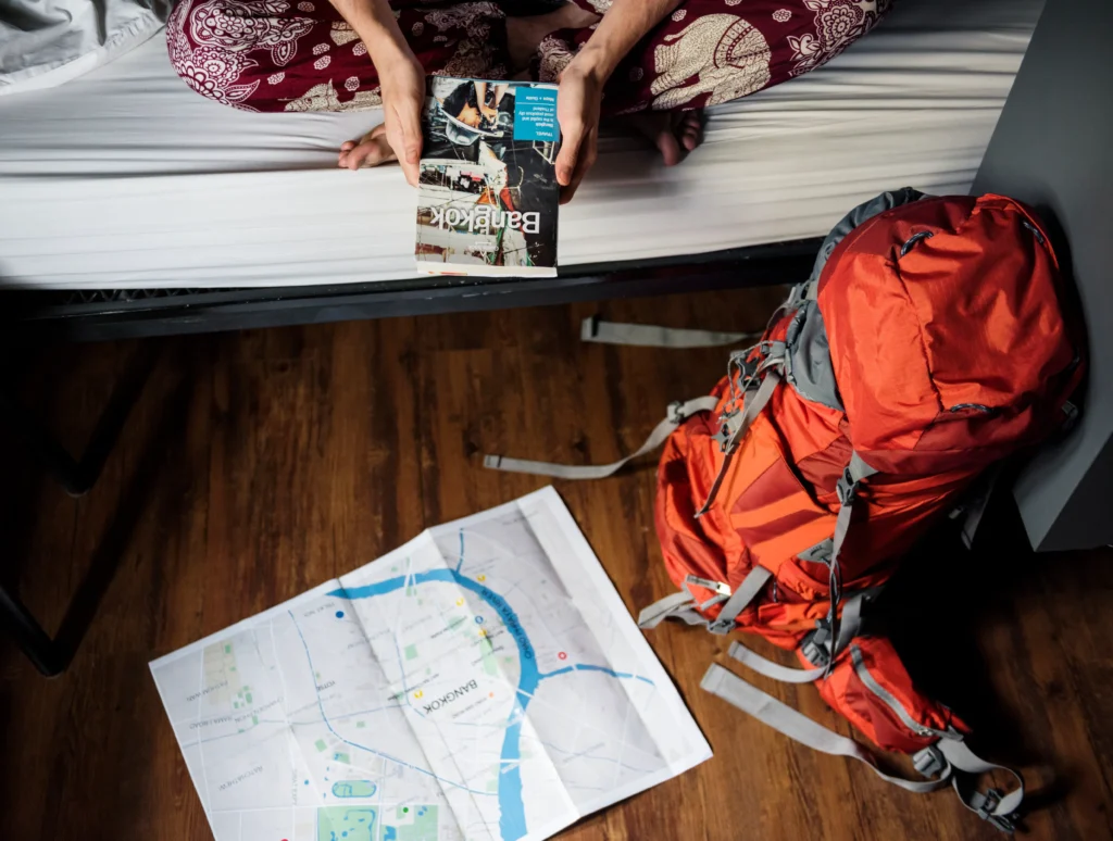 Persona en cama con guía de Bangkok, junto a su mochila para viajar naranja y un mapa en el suelo de madera.