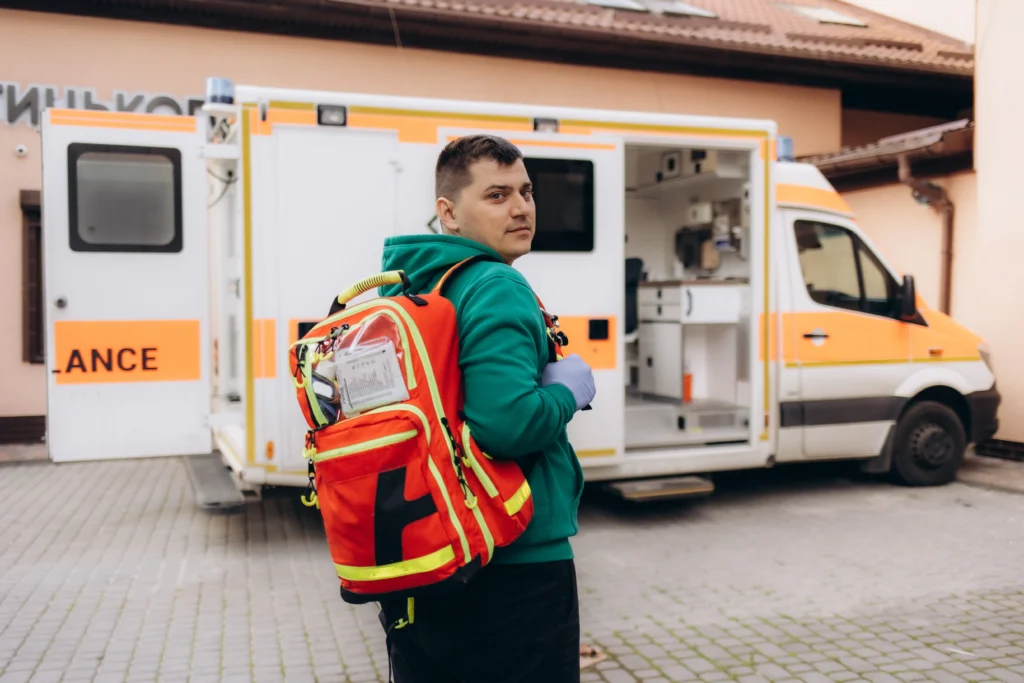 Un rescatista con mochilas de rescate en la espalda se gira para mirar a la cámara delante de una ambulancia abierta.